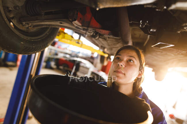 Mixed race female car mechanic wearing overalls, replacing car oil. independent business owner at car servicing garage. — Stock Photo