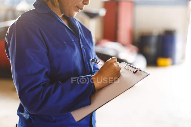 Midsection of mixed race female car mechanic wearing overalls, making notes. independent business owner at car servicing garage. — Stock Photo
