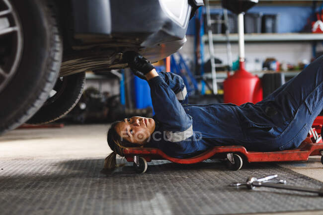 Mécanicien automobile mixte féminin en salopette, allongé sur une planche, vérifiant la voiture. propriétaire d'entreprise indépendant au garage d'entretien de voiture. — Photo de stock