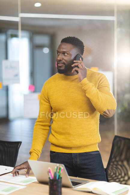 Sonriente hombre afroamericano negocio creativo de pie en el escritorio y el uso de teléfonos inteligentes. empresarios creativos independientes que trabajan en una oficina moderna. - foto de stock