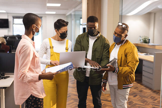 Diverse männliche und weibliche Kollegen tragen Gesichtsmasken und arbeiten mit Laptop zusammen. Arbeit im kreativen Geschäft in einem modernen Büro während der Coronavirus-Pandemie. — Stockfoto