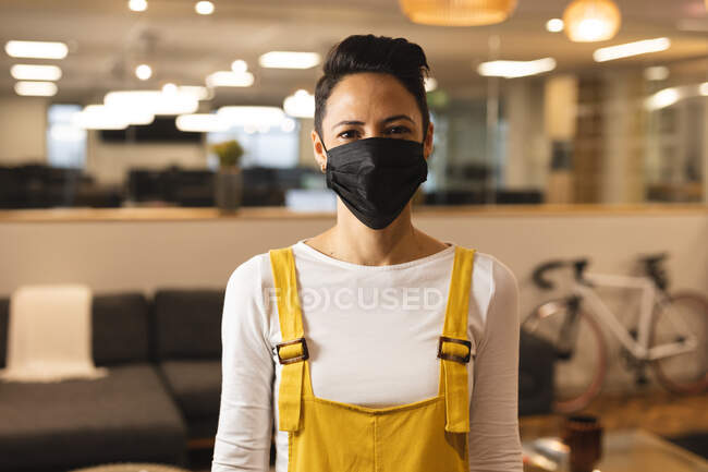 Retrato de mujer de raza mixta creativa usando mascarilla en el trabajo, mirando a la cámara. trabajar en negocios creativos en una oficina moderna durante la pandemia de coronavirus. - foto de stock