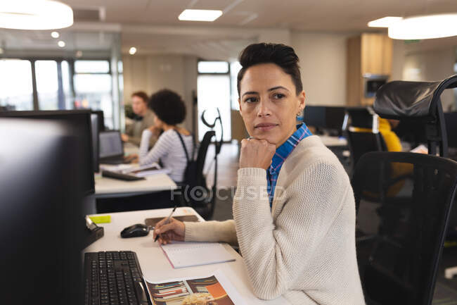 Carrera mixta femenina creativa en el trabajo, sentada en el escritorio, utilizando el ordenador. trabajar en un negocio creativo en una oficina moderna. - foto de stock