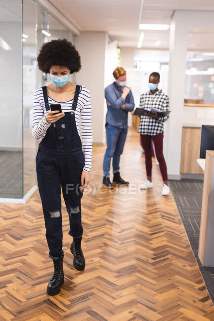 Разнообразные коллеги мужского и женского пола в масках для лица, работающие вместе с помощью смартфона. работа в творческом бизнесе в современном офисе во время пандемии коронавируса. — стоковое фото