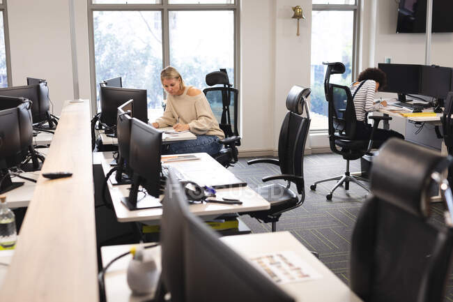 Diversas compañeras en el trabajo, sentadas en escritorios, tomando notas. trabajar en un negocio creativo en una oficina moderna. - foto de stock