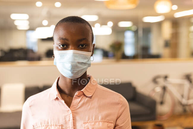 Ritratto di donna afroamericana creativa che indossa una maschera facciale al lavoro, guardando alla macchina fotografica. lavorare nel business creativo in un ufficio moderno durante la pandemia di coronavirus. — Foto stock
