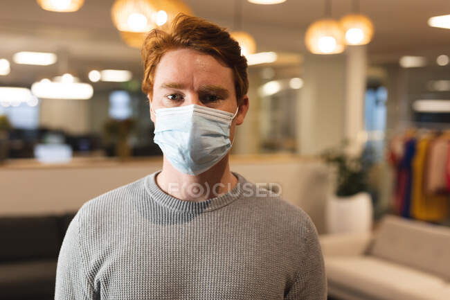 Ritratto di maschio caucasico creativo che indossa una maschera facciale al lavoro, guardando alla macchina fotografica. lavorare nel business creativo in un ufficio moderno durante la pandemia di coronavirus. — Foto stock