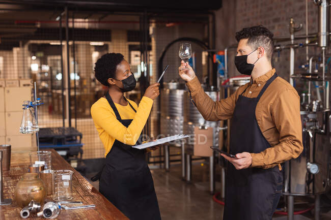 Diverse man and woman wearing face masks checking gin product in glass at gin distillery. alcohol production and filtration concept — Stock Photo