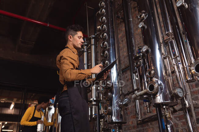 Белый мужчина в фартуке держит планшет, проверяющий оборудование на заводе. Концепция производства и фильтрации алкоголя — стоковое фото