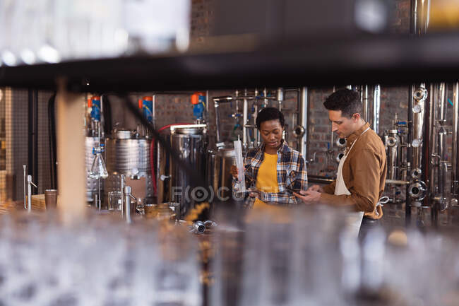 Homem caucasiano e mulher afro-americana a verificar o produto gin em frasco na destilaria de gin. conceito de produção e filtração de álcool — Fotografia de Stock