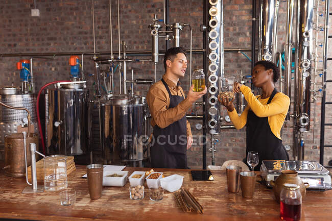 Hombre diverso mujer sosteniendo un frasco de ingredientes para la producción de ginebra en la destilería de ginebra. concepto de producción y filtración de alcohol. - foto de stock