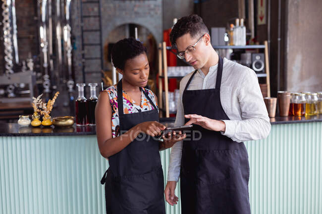 Homme caucasien et femme afro-américaine portant des tabliers utilisant une tablette numérique à la distillerie de gin. artisanat indépendant gin distillerie concept d'entreprise — Photo de stock