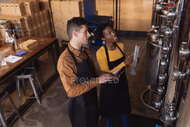 Diverse man and woman holding digital tablet and flask checking equipment at gin distillery. alcohol production and filtration concept — Stock Photo
