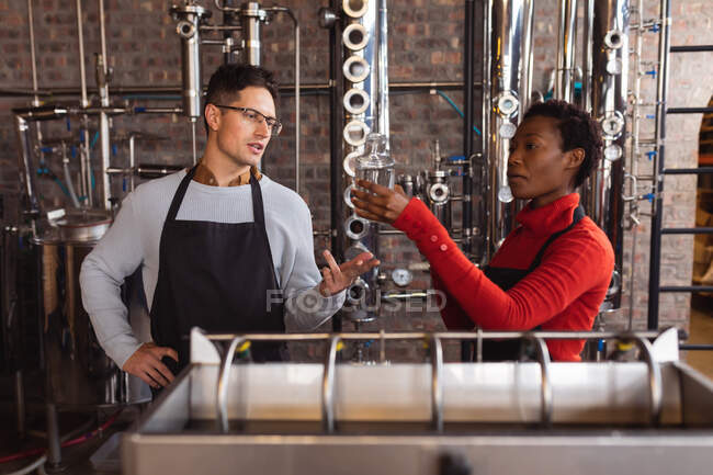 Homem caucasiano e mulher afro-americana a verificar garrafa de gin em vidro na destilaria de gin. conceito de produção e filtração de álcool — Fotografia de Stock