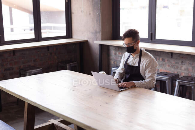 African american woman wearing apron and face mask using laptop at gin distillery. independent craft gin distillery business during covid-19 pandemic concept — Stock Photo
