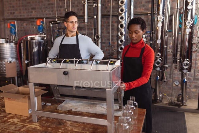 Kaukasischer Mann und afrikanisch-amerikanische Frau mit Maschinen in der Gin-Brennerei. Alkoholproduktion und Filtrationskonzept — Stockfoto