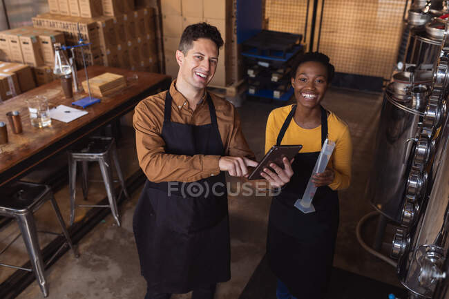Portrait of diverse man and woman holding digital tablet and flask smiling at gin distillery. alcohol production and filtration concept — Stock Photo