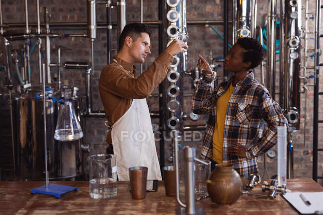 Кавказький чоловік і афро-американська жінка перевіряють, чи не виготовляють джин у склі під час винокурні. Виробництво алкоголю і фільтрація. — стокове фото