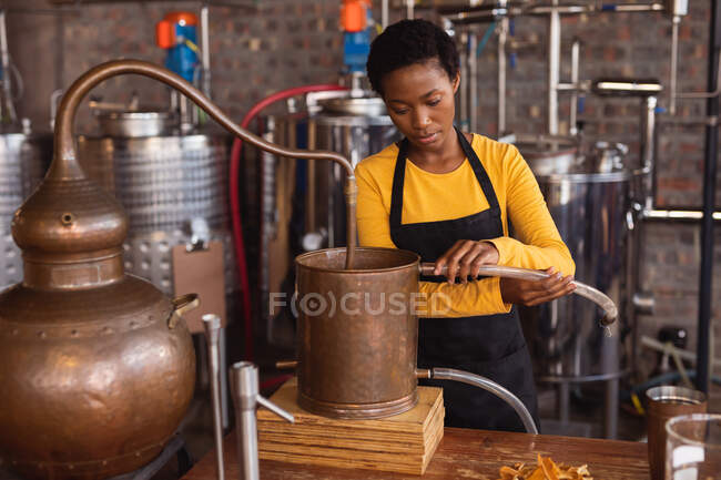 Donna afroamericana che collega il tubo alle apparecchiature di distillazione della distilleria di gin. produzione di alcol e concetto di filtrazione — Foto stock
