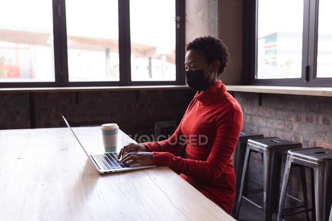 African american woman wearing a face mask using laptop at gin distillery. independent craft gin distillery business during covid-19 pandemic concept — Stock Photo