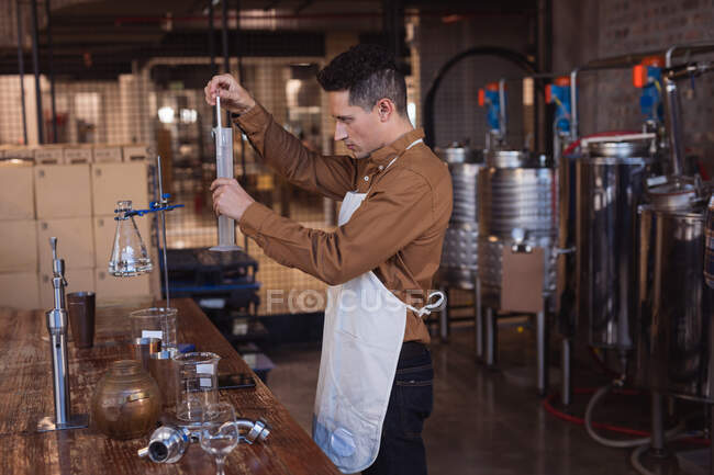 Кавказький чоловік у фартусі перевіряє продукт джину у колбі на винокурні джину. Виробництво алкоголю і фільтрація. — стокове фото