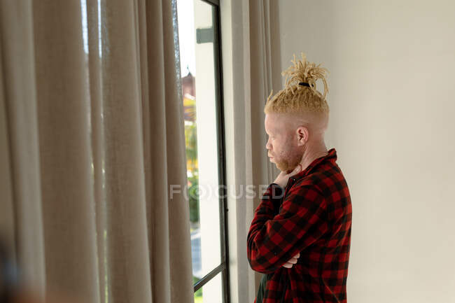 Pensativo albino hombre afroamericano con rastas mirando por la ventana. trabajo remoto utilizando tecnología en el hogar. - foto de stock