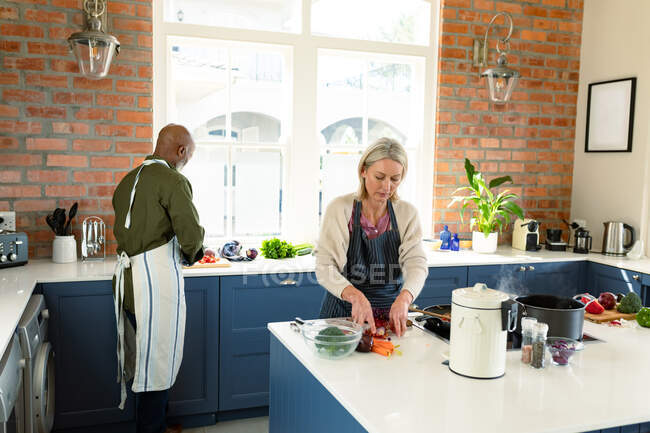 Feliz casal diversificado sênior na cozinha vestindo aventais, cozinhar juntos. estilo de vida saudável e ativo em casa. — Fotografia de Stock
