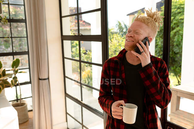 Улыбающийся альбинос африканский американец с дредами, использующий смартфон и пьющий кофе. удаленная работа с использованием технологии на дому. — стоковое фото