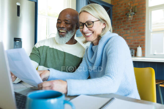 Heureux couple diversifié senior dans la cuisine assis à table, en utilisant un ordinateur portable. mode de vie à la retraite, à la maison avec la technologie. — Photo de stock