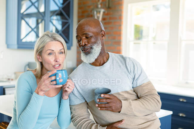 Happy senior diverse couple in kitchen sitting at countertop, drinking coffee. retirement lifestyle, spending time at home. — Stock Photo