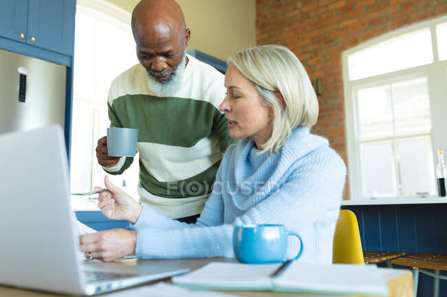 Sforzato anziani coppia diversificata in cucina seduto a tavola, utilizzando il computer portatile. stile di vita di pensione, a casa con la tecnologia. — Foto stock