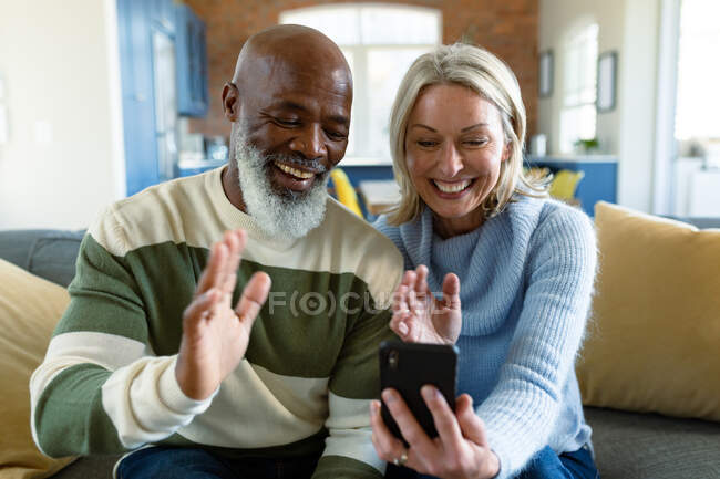 Felice anziano coppia diversificata in soggiorno seduto sul divano, utilizzando smartphone. stile di vita di pensione, a casa con la tecnologia. — Foto stock