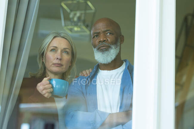 Worried senior diverse couple looking through window and embracing. retirement lifestyle, spending time at home. — Stock Photo