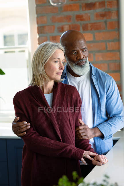 Preocupada pareja de ancianos diversos en la cocina apoyada en la encimera, abrazando y mirando hacia otro lado. estilo de vida de jubilación, pasar tiempo en casa. - foto de stock