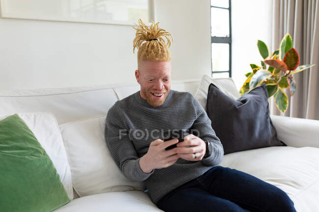 Щасливий альбіно-афроамериканський чоловік у вітальні за допомогою смартфона. відпочинок за допомогою технології, відпочинок вдома . — стокове фото