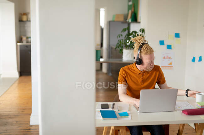 Albino hombre afroamericano con rastas trabajando desde casa y haciendo videollamadas en el portátil. trabajo remoto utilizando tecnología en el hogar. - foto de stock