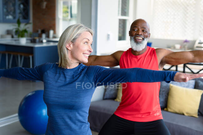 Feliz pareja mayor diversa en ropa de ejercicio practicando yoga juntos, estirándose. estilo de vida saludable y activo de jubilación en el hogar. - foto de stock