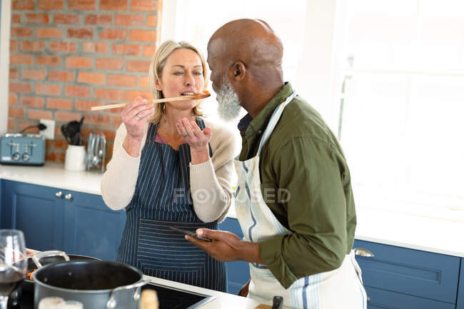 Happy senior diverse couple in kitchen wearing aprons, cooking together, trying food. healthy, active retirement lifestyle at home. — Stock Photo