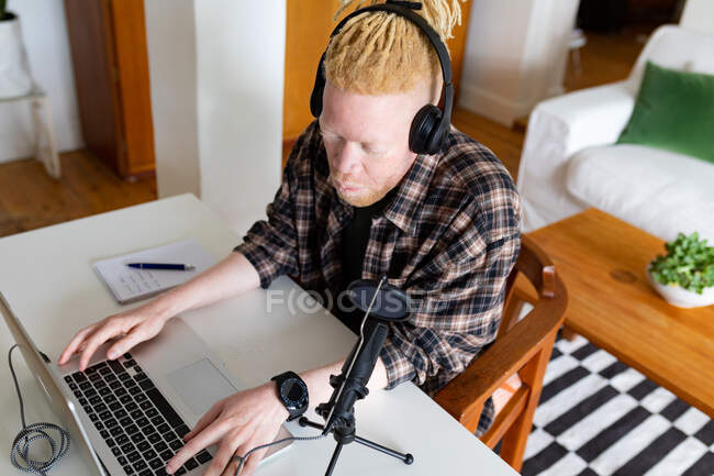 Glücklicher Albino-Afrikaner mit Dreadlocks, der von zu Hause aus arbeitet und Podcasts macht. Fernbedienung mit Technologie zu Hause. — Stockfoto