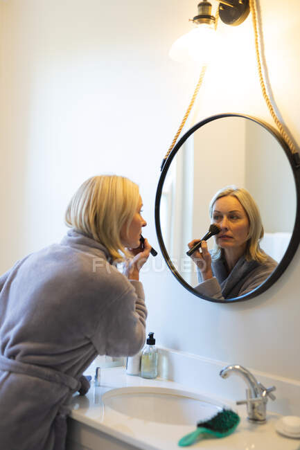 Mulher caucasiana sênior feliz na casa de banho, olhando para o espelho, colocando maquiagem. estilo de vida da aposentadoria, passar tempo em casa. — Fotografia de Stock
