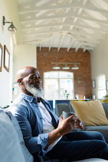 Feliz homem afro-americano sênior na sala de estar sentado no sofá, segurando caneca. estilo de vida da aposentadoria, passar tempo em casa. — Fotografia de Stock