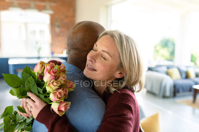 Felice anziano coppia diversificata in soggiorno abbracciando, tenendo fiori. stile di vita di pensione, trascorrere del tempo a casa. — Foto stock