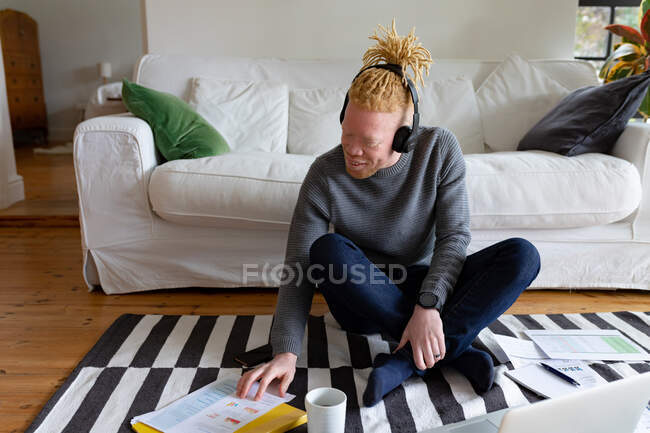 Feliz albino hombre afroamericano sentado en el suelo y trabajando desde casa usando el ordenador portátil. trabajo remoto utilizando tecnología en el hogar. - foto de stock
