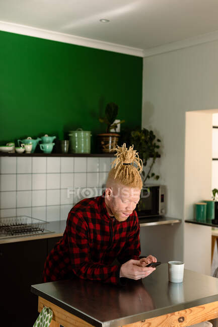 Sorrindo albino homem americano africano com dreadlocks usando smartphone e beber café. trabalho remoto usando a tecnologia em casa. — Fotografia de Stock