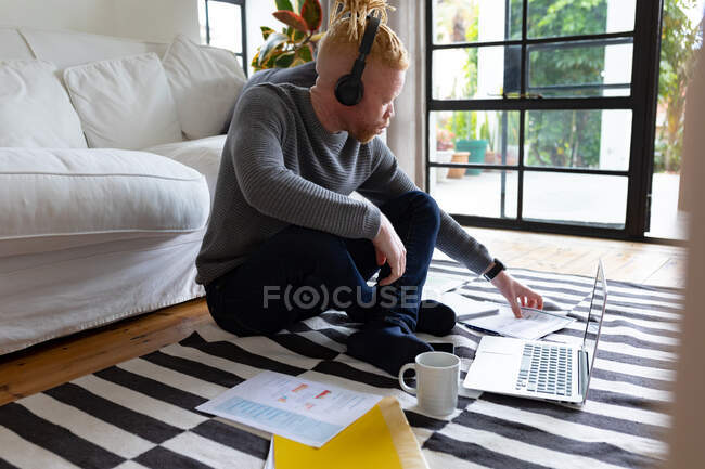 Albino hombre afroamericano sentado en el suelo y trabajando desde casa usando el ordenador portátil. trabajo remoto utilizando tecnología en el hogar. - foto de stock