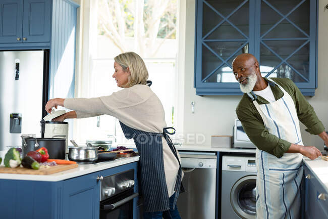 Felice anziano coppia diversificata in cucina indossando grembiuli, cucinare insieme. stile di vita sano e attivo pensionamento a casa. — Foto stock