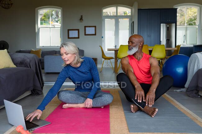 Casal diverso sênior feliz em roupas de exercício praticando ioga juntos, usando laptop. estilo de vida saudável e ativo em casa. — Fotografia de Stock