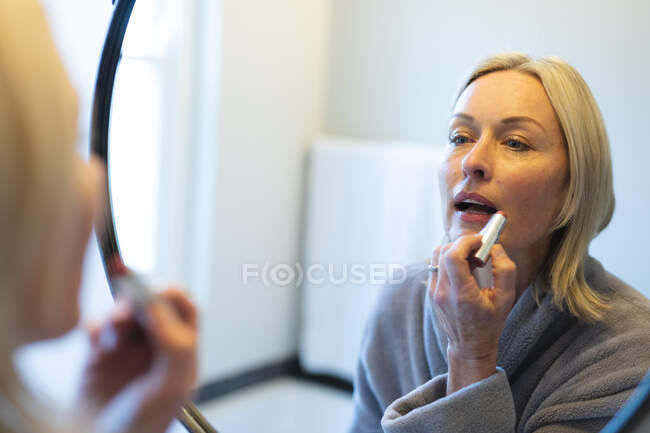 Glückliche ältere kaukasische Frau im Badezimmer, schaut in den Spiegel und setzt Lippenstift auf. Lebensstil im Ruhestand, Zeit zu Hause verbringen. — Stockfoto