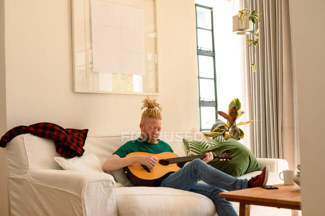 Albino uomo afroamericano con dreadlocks in salotto a suonare la chitarra. tempo libero, relax a casa. — Foto stock