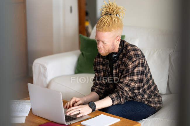Hombre afroamericano albino con rastas trabajando desde casa y usando laptop. trabajo remoto utilizando tecnología en el hogar. - foto de stock